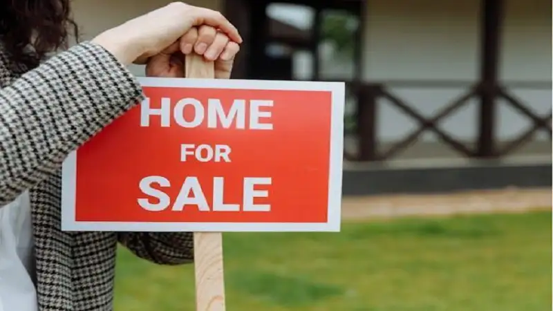 Person holding a red “Home For Sale” sign in front of a house